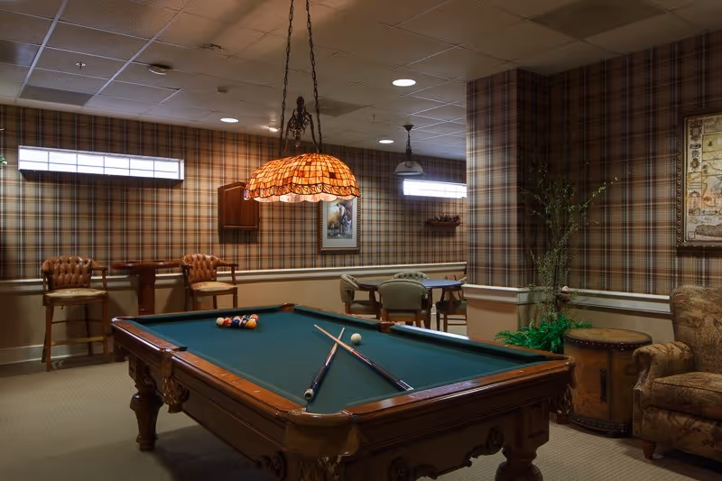 A cozy game room with a pool table in the center, two pool cues crossed on the table, and a rack of billiard balls. The room features plaid wallpaper, a hanging stained glass lamp above the pool table, two high chairs with a small table against the wall, and a seating area with an armchair and a round side table. There is also a framed picture on the wall and some plants adding greenery to the space.