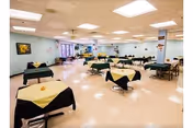 A spacious dining room with multiple tables covered in black tablecloths and yellow napkins. The room has light-colored walls and ceiling with fluorescent lighting. There are windows along one side and some decorations on the walls.