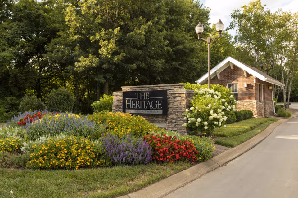 Entrance sign for The Heritage at Brentwood surrounded by colorful flowers and greenery, with a small brick building and street lamps in the background.