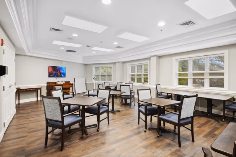 A bright and spacious dining room with multiple square wooden tables and cushioned chairs arranged neatly. The room has large windows with white blinds allowing natural light to fill the space. The floor is wooden, and the ceiling features recessed lighting and decorative panels. A piano and a flat-screen TV mounted on the wall are visible in the background.