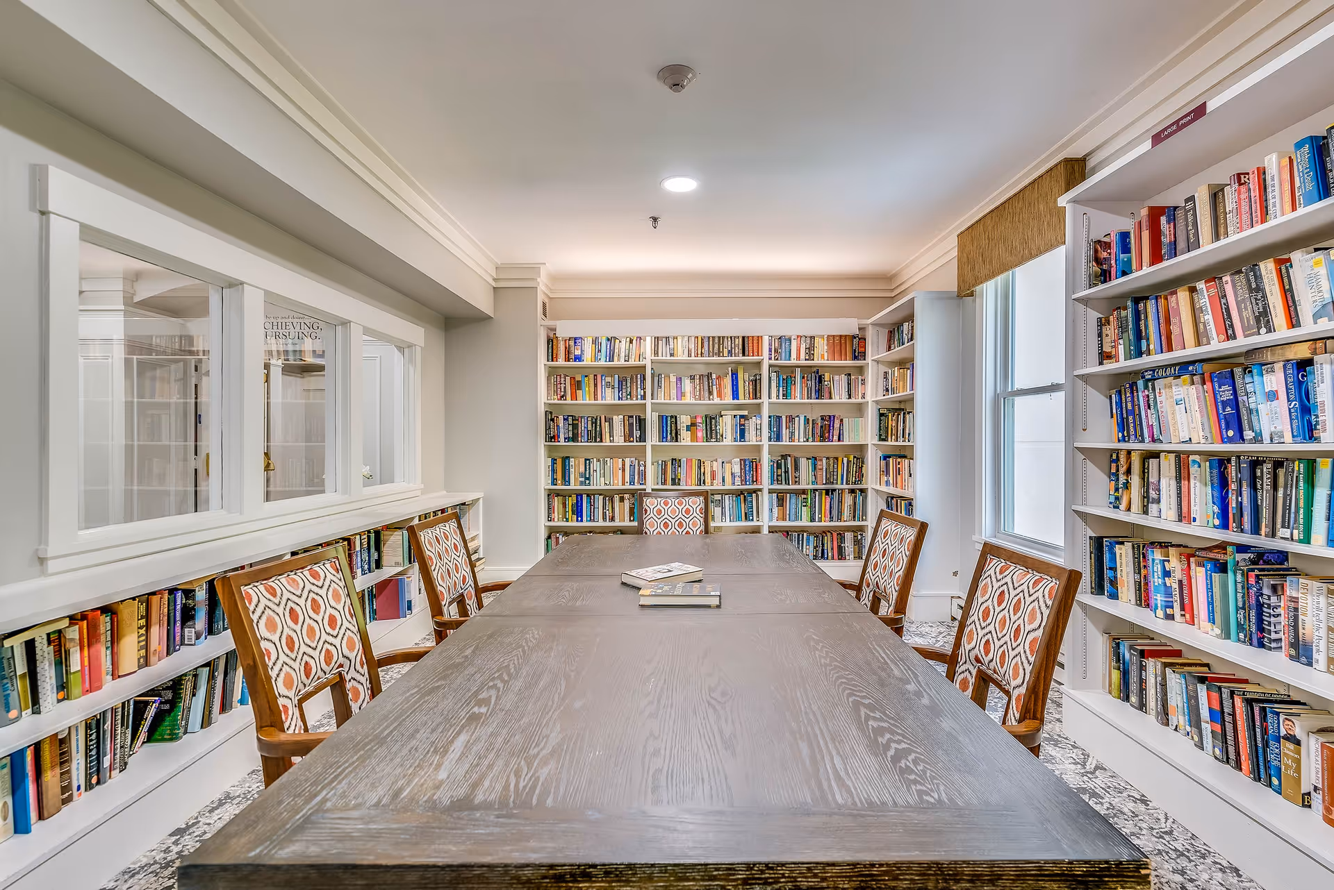 Long wooden table surrounded by patterned chairs in a bright library room lined with bookshelves.