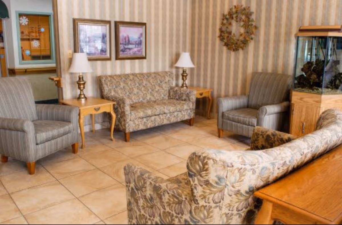 Cozy assisted-living lounge with patterned sofas and armchairs, side tables with lamps, framed art, and an aquarium.