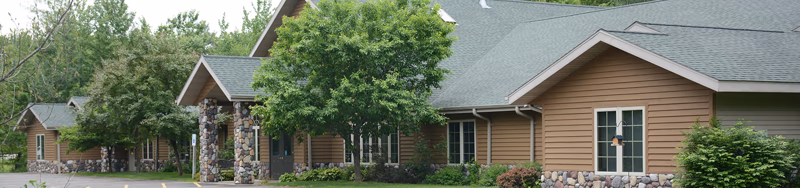 Exterior view of a single-story assisted living facility building with brown siding, stone accents, multiple windows, and a green roof surrounded by trees and shrubs.