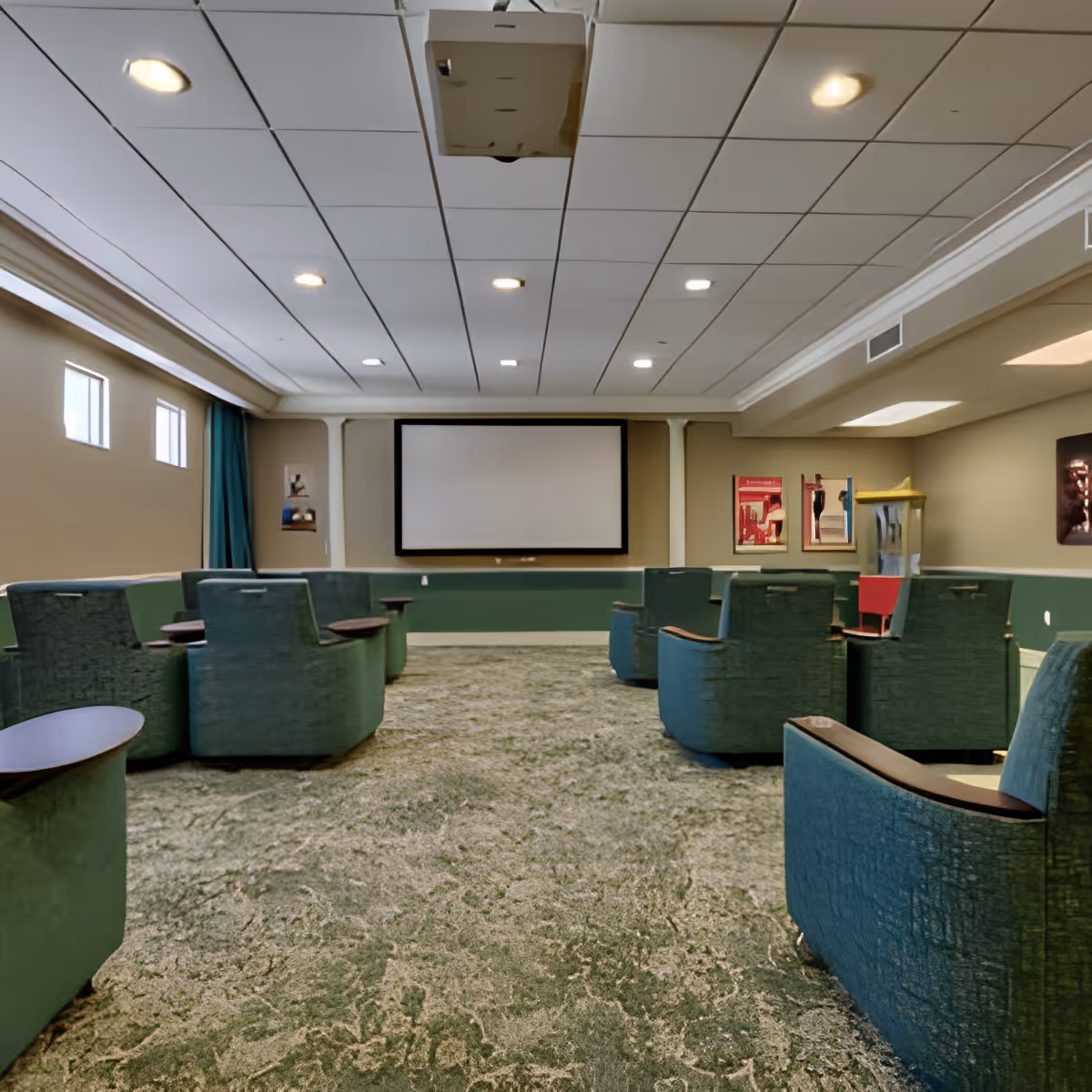 A small theater room with green upholstered chairs arranged in rows facing a large blank projection screen on the wall. The room has a patterned carpet, beige walls, and a popcorn machine in the corner. There are small windows near the ceiling on the left side and recessed lighting in the ceiling.