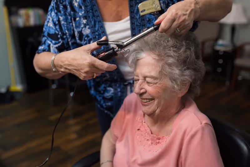 An elderly woman with gray curly hair is sitting in a chair smiling with her eyes closed while another person styles her hair using a curling iron in an indoor setting.