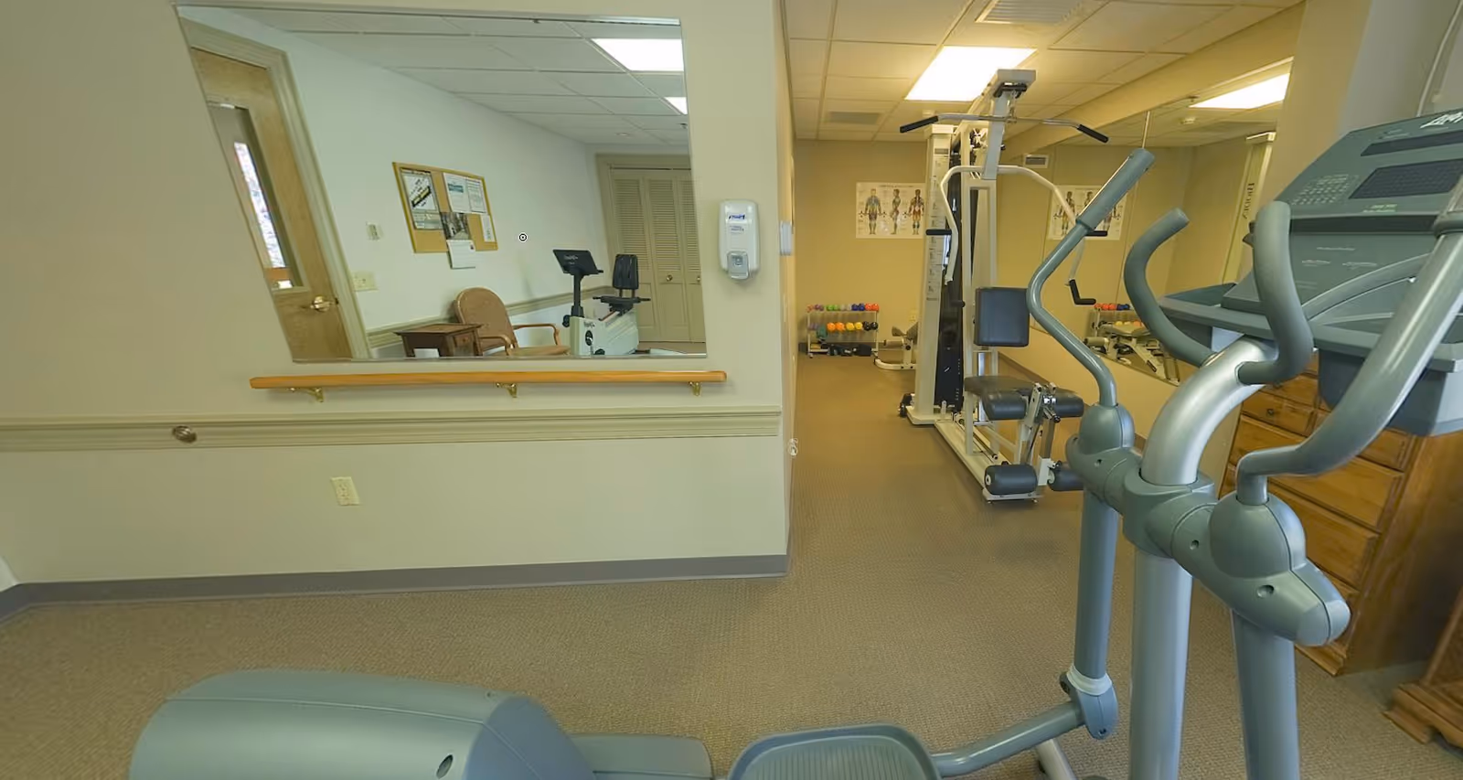 Interior view of a fitness room in a senior living facility featuring exercise equipment including an elliptical machine, a multi-station gym machine, and a stationary bike. The room has beige walls, carpeted floor, a large mirror on one wall, and anatomical posters. There is a hand sanitizer dispenser mounted on the wall and a bulletin board with notices.