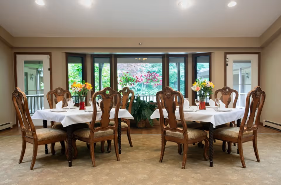 A dining room with two tables covered with white tablecloths, each surrounded by four ornate wooden chairs with cushioned seats. Each table has a vase with colorful flowers and place settings with napkins and glasses. Large windows and glass doors in the background provide a view of greenery outside.