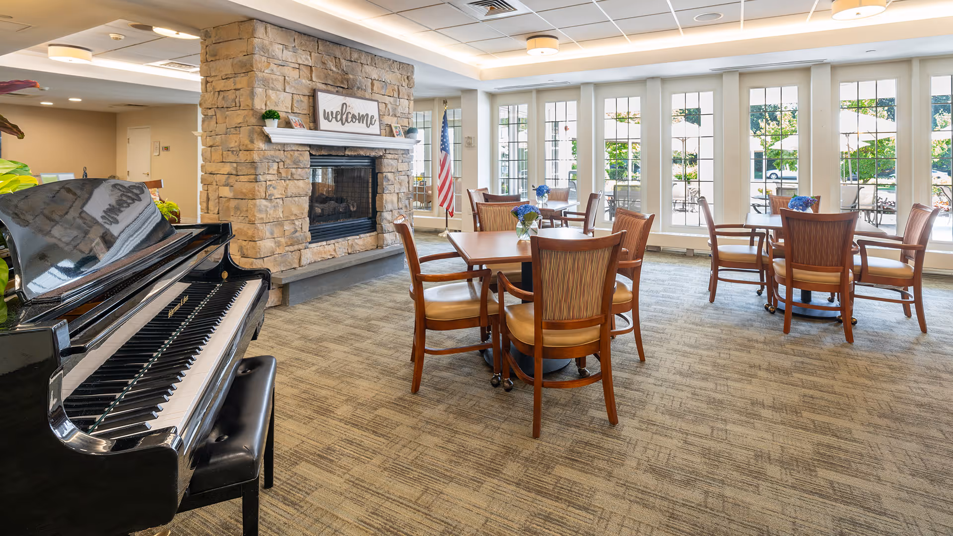 A bright and welcoming common area in a senior living facility featuring a black grand piano on the left, a stone fireplace with a 'welcome' sign above it, several wooden tables with chairs arranged around them, and large windows letting in natural light with a view of an outdoor patio area.