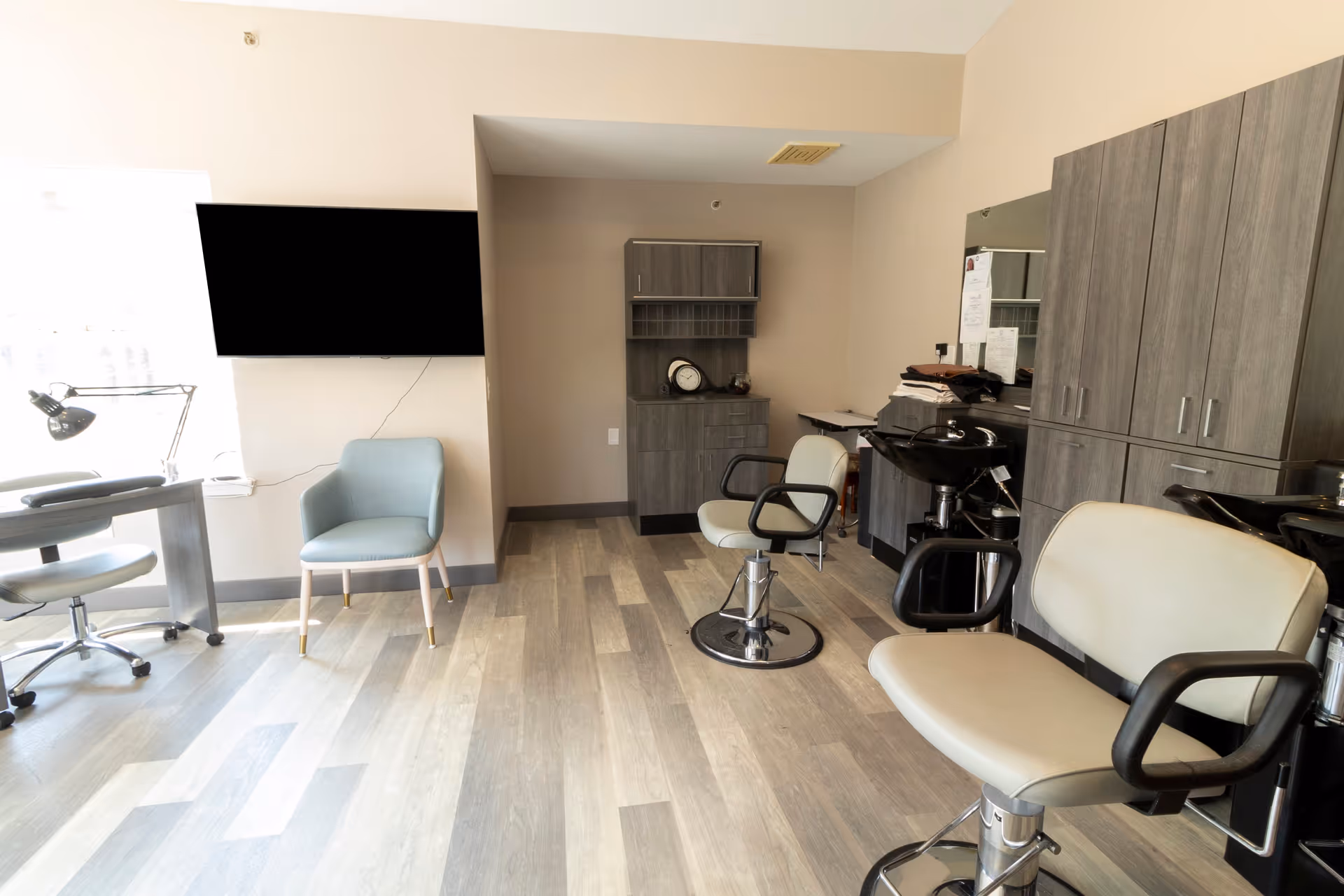 Interior of a salon room with two salon chairs in front of hair washing stations, a blue armchair near a window, a wall-mounted TV, a desk with a lamp, and wooden cabinets along the wall.