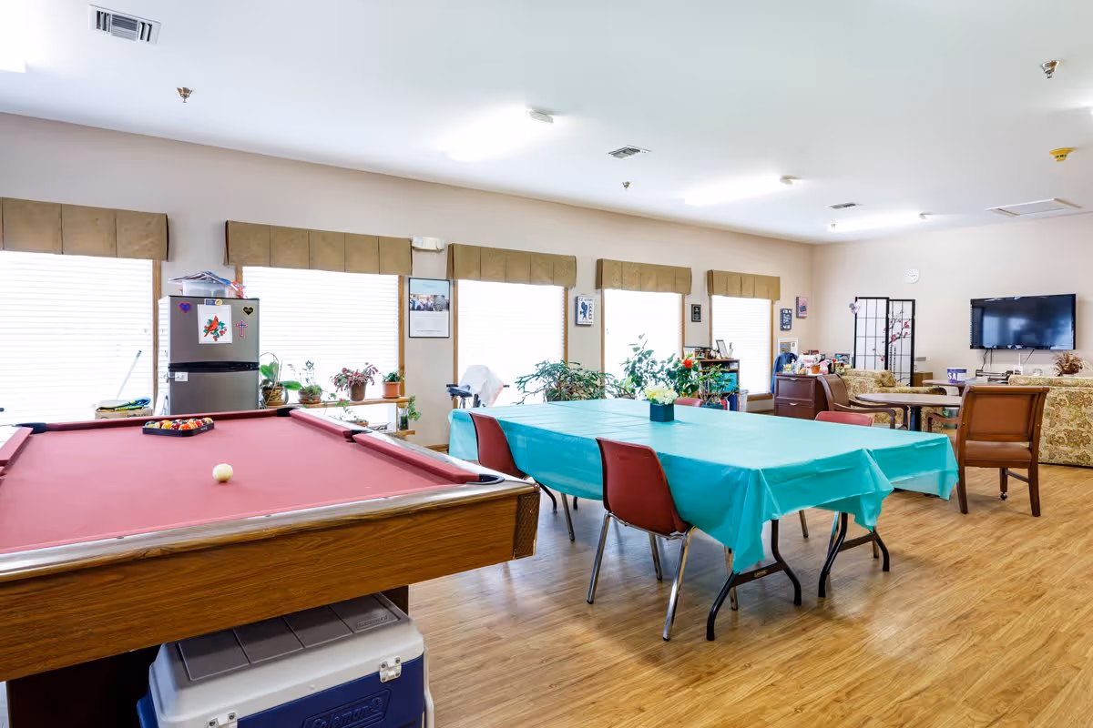 A bright and spacious common room with a red pool table in the foreground, a table covered with a teal tablecloth surrounded by chairs, several potted plants on the windowsills, a refrigerator with magnets, and a seating area with couches and a wall-mounted TV in the background.