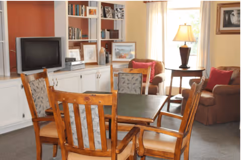 A cozy living room area with a wooden table and four chairs in the foreground. In the background, there is a built-in white bookshelf with various books and decorative items, a television, and two armchairs with red cushions near a window with light curtains. A table lamp is placed on a side table between the armchairs.