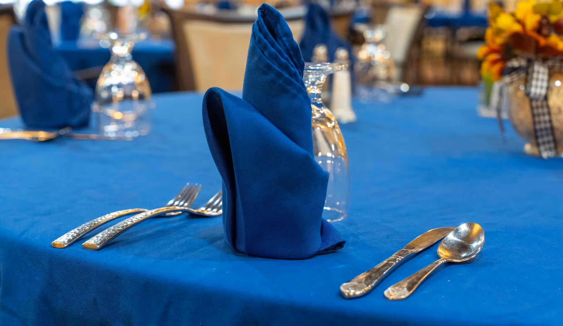 Close-up of a dining table set with a blue tablecloth, blue folded napkin, upside-down wine glass, two forks on the left, and a knife and spoon on the right. In the background, there are more tables with similar settings and a floral centerpiece with orange and yellow flowers tied with a checkered ribbon.