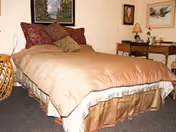 A neatly made bed with a beige and gold comforter and multiple decorative pillows in a bedroom. The room has carpeted flooring, a wooden desk with a lamp and flowers, and framed artwork on the walls.