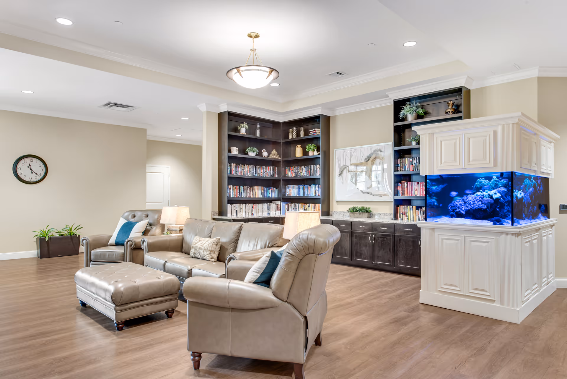 A bright and spacious living room area with beige leather sofas and an ottoman, two table lamps, a large built-in dark wood bookshelf filled with books and decorative plants, a large aquarium with blue lighting, a framed horse artwork on the wall, a clock, and wooden flooring.