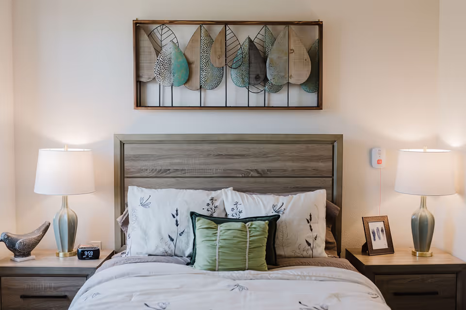 A neatly made bed with a wooden headboard, white and floral-patterned pillows, and a green decorative pillow in the center. On each side of the bed are matching wooden nightstands with table lamps, a decorative bird figurine on the left nightstand, and a framed picture on the right nightstand. Above the bed is a wall decoration featuring metal and wooden leaves.