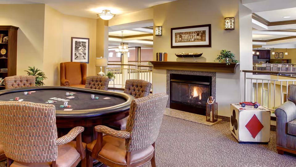 A cozy common area in a senior living facility featuring a poker table with chairs around it, a lit fireplace with a mantel decorated with books and a bowl, framed pictures on the walls, and comfortable seating including an armchair and a cube-shaped ottoman with card suit symbols. The space has warm lighting and overlooks a railing with more seating areas visible in the background.