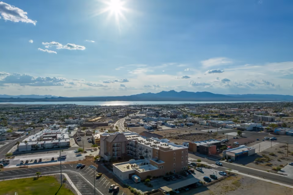 Aerial view of a residential and commercial area with a multi-story building in the foreground, roads, parked cars, and a large body of water with mountains in the background under a partly cloudy sky with the sun shining.