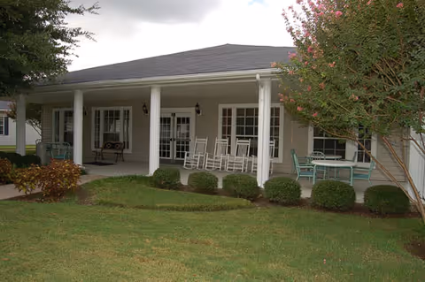 Covered porch area of a building with white columns, several rocking chairs, a bench, and a table with chairs. The porch overlooks a well-maintained lawn with shrubs and trees.