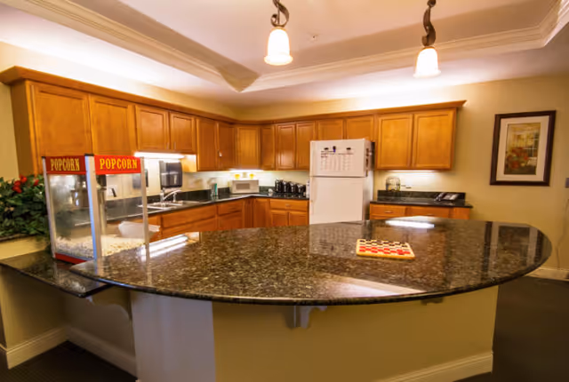 A kitchen area with wooden cabinets, a white refrigerator, microwave, toaster, and a popcorn machine on a large curved granite countertop. There is a framed picture on the wall and some plants in the corner.