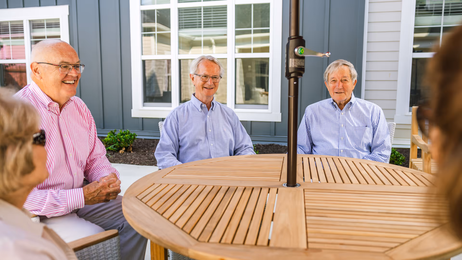 A group of elderly people sitting around a wooden outdoor table with an umbrella pole in the center, smiling and engaging in conversation outside a building with gray siding and white-framed windows.