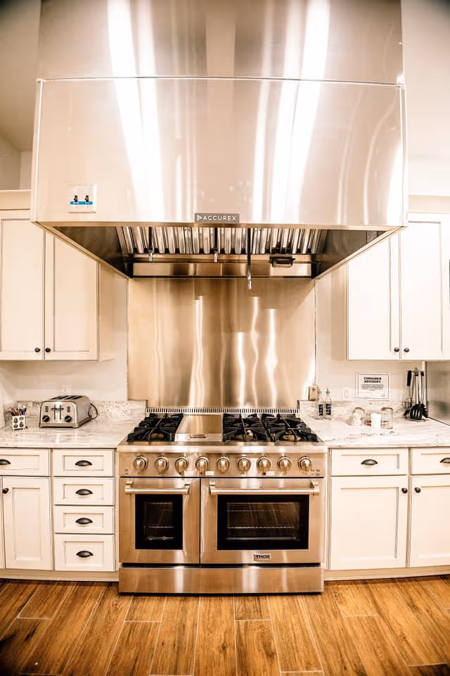 Modern kitchen with a large stainless steel stove and oven, a stainless steel range hood above, white cabinets with black handles, a toaster on the left countertop, and various kitchen utensils on the right countertop. The floor is wooden.
