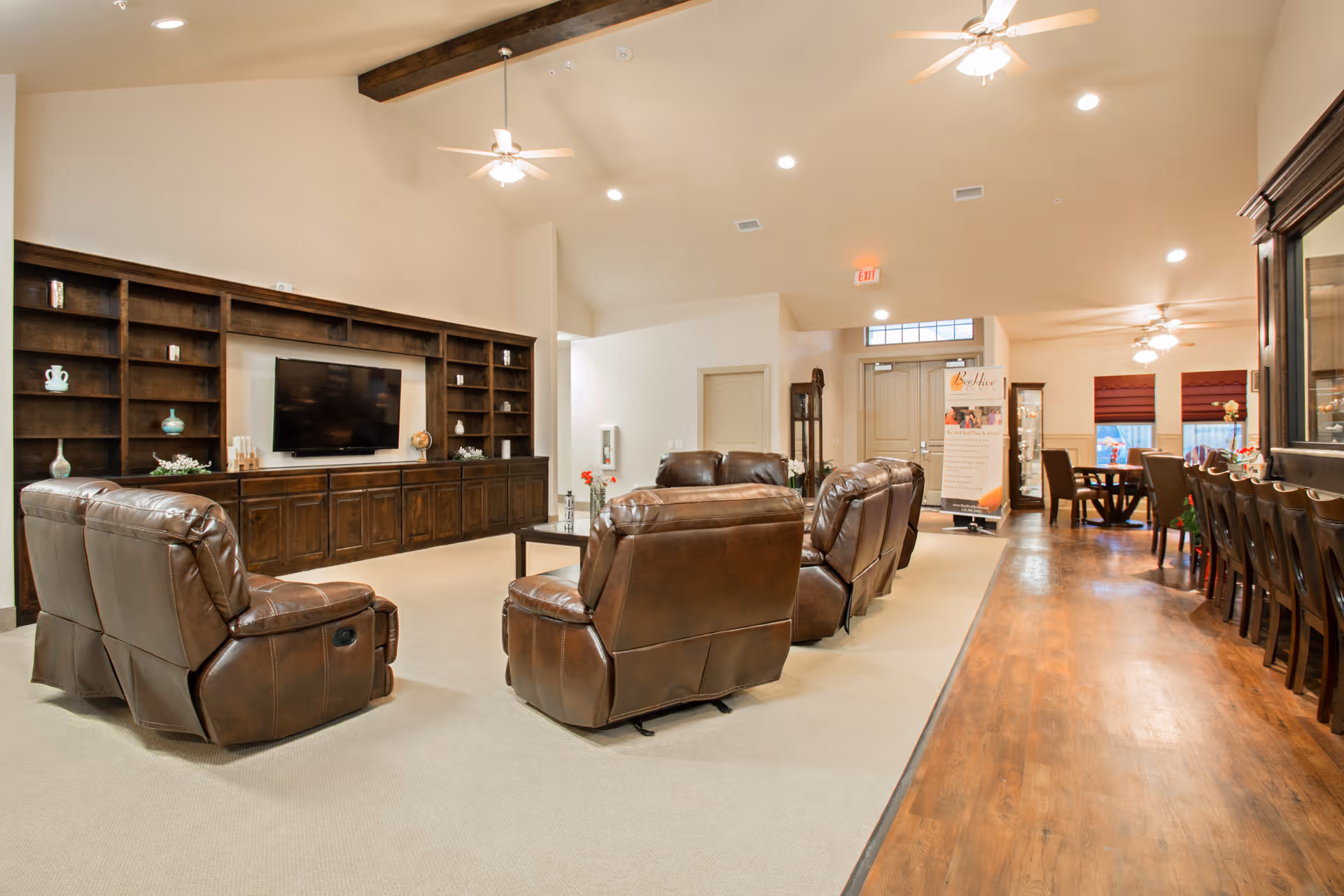 A spacious living room area in a senior living facility featuring four brown leather recliners arranged around a coffee table. The room has a large built-in wooden shelving unit with a flat-screen TV mounted in the center. The ceiling is vaulted with exposed wooden beams and ceiling fans. To the right, there is a long wooden dining table with chairs and a smaller round table with chairs near windows with red blinds. The flooring is a combination of carpet and wood, and the room is well-lit with recessed lighting.