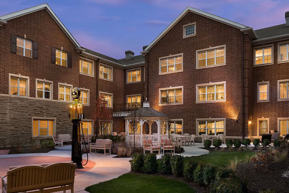 Evening view of the outdoor courtyard at Cedarbrook Of Bloomfield Hills, featuring a brick building with multiple lit windows, a white gazebo, outdoor seating with chairs and benches, landscaped greenery, and a vintage-style gas pump.