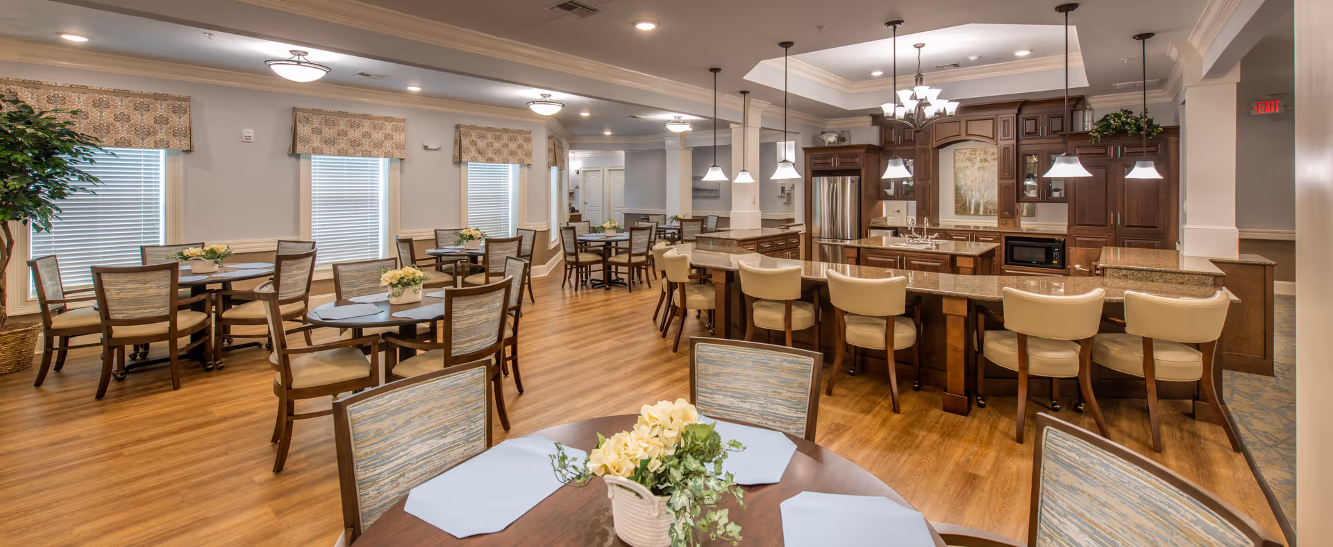 A spacious dining area in a senior living facility featuring multiple round tables with chairs, each table decorated with a small floral centerpiece. The room has large windows with patterned valances, wood flooring, and a kitchen area with a long counter and bar stools. Pendant lights hang above the counter, and the kitchen has wooden cabinetry and stainless steel appliances.