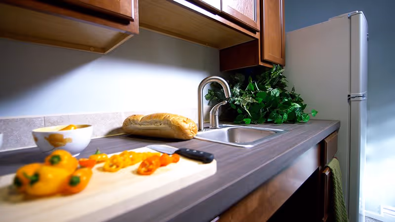 A kitchen countertop with a cutting board holding sliced yellow and orange bell peppers and a knife. There is a loaf of bread and a bowl with fruit on the counter near a stainless steel sink with a faucet. Wooden cabinets are above and below the counter, and a white refrigerator is visible in the background. A green leafy plant is placed near the sink.