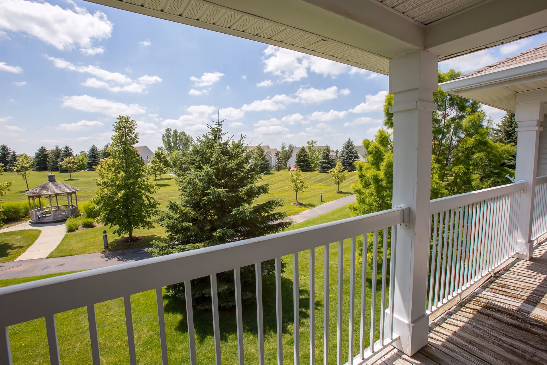 View from a covered balcony with white railing overlooking a green lawn with trees, a gazebo, and a paved walkway under a partly cloudy sky.