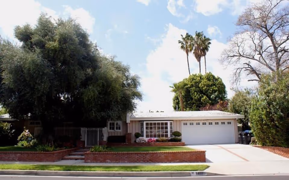 Front exterior view of a single-story residential building with a large tree on the left side, a well-maintained lawn, brick planters, and a two-car garage. The sky is partly cloudy with some palm trees visible in the background.