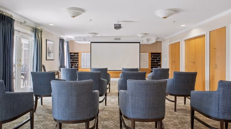 A small conference or presentation room with rows of blue upholstered chairs facing a large white projection screen. The room has beige walls, wooden doors on the right side, and large windows with dark curtains on the left side letting in natural light.