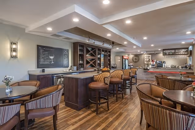 Interior view of a dining area with wooden floors, round tables with striped cushioned chairs, a bar counter with high stools, and a chalkboard menu on the wall. The space is well-lit with recessed ceiling lights and has a cozy, inviting atmosphere.