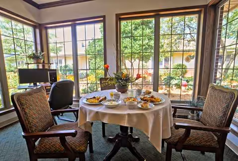 Round dining table with a white tablecloth and plates of food by sunlit windows.