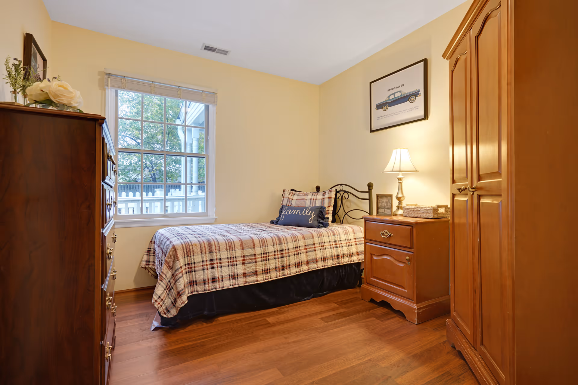 A cozy bedroom with a single bed covered in a plaid blanket and a pillow with the word 'family'. The room has wooden furniture including a dresser, nightstand with a lamp, and a wardrobe. A large window shows a white picket fence and trees outside. The walls are painted light yellow and there is a framed picture of a vintage car above the bed.