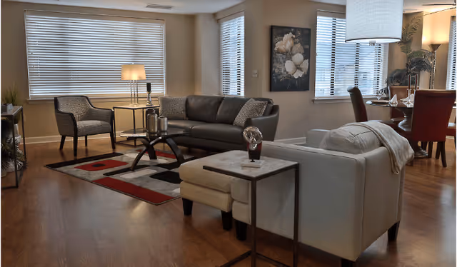 A cozy living room area with a gray sofa, a white armchair with a throw blanket, a patterned rug, and a small side table with decorative items. There is a dining area with a table and red chairs in the background, and large windows with blinds letting in natural light.