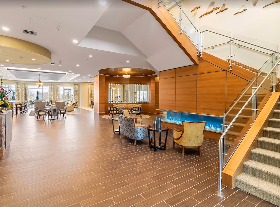 A spacious and modern interior of a senior living facility featuring a seating area with armchairs and small tables near a contemporary fireplace under a staircase with glass and metal railing. The room has wood-paneled walls, large windows with curtains, and additional seating and dining tables in the background.