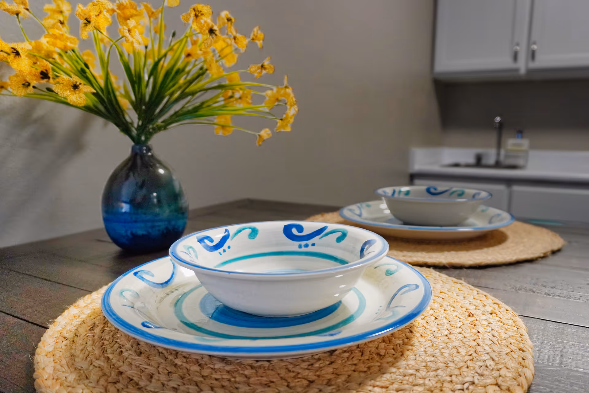 Close-up view of a dining table set with two place settings, each consisting of a decorative bowl on a matching plate placed on woven placemats. A blue vase with yellow flowers is on the table, and a kitchen counter with a sink and cabinets is visible in the background.