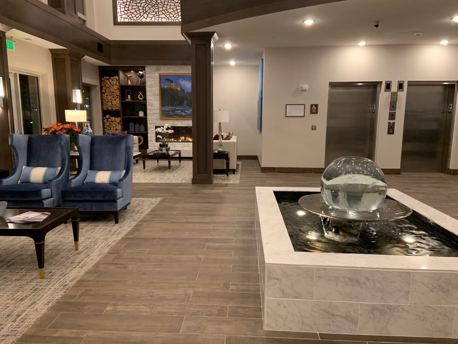Interior view of a senior living facility lobby featuring a modern water fountain with a glass sphere centerpiece, two blue upholstered armchairs with pillows, a coffee table with magazines, a fireplace with a painting above it, and two elevators in the background.