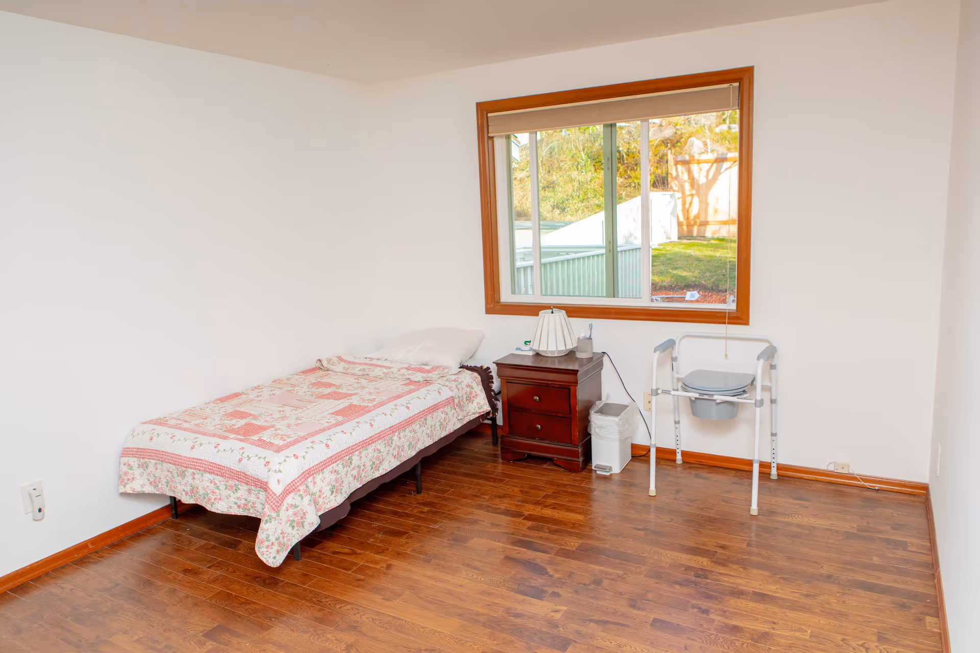 A simple bedroom with a single bed covered with a floral quilt, a wooden nightstand with a lamp and some items on it, a trash bin, and a portable commode chair near a large window with a view of a grassy outdoor area.