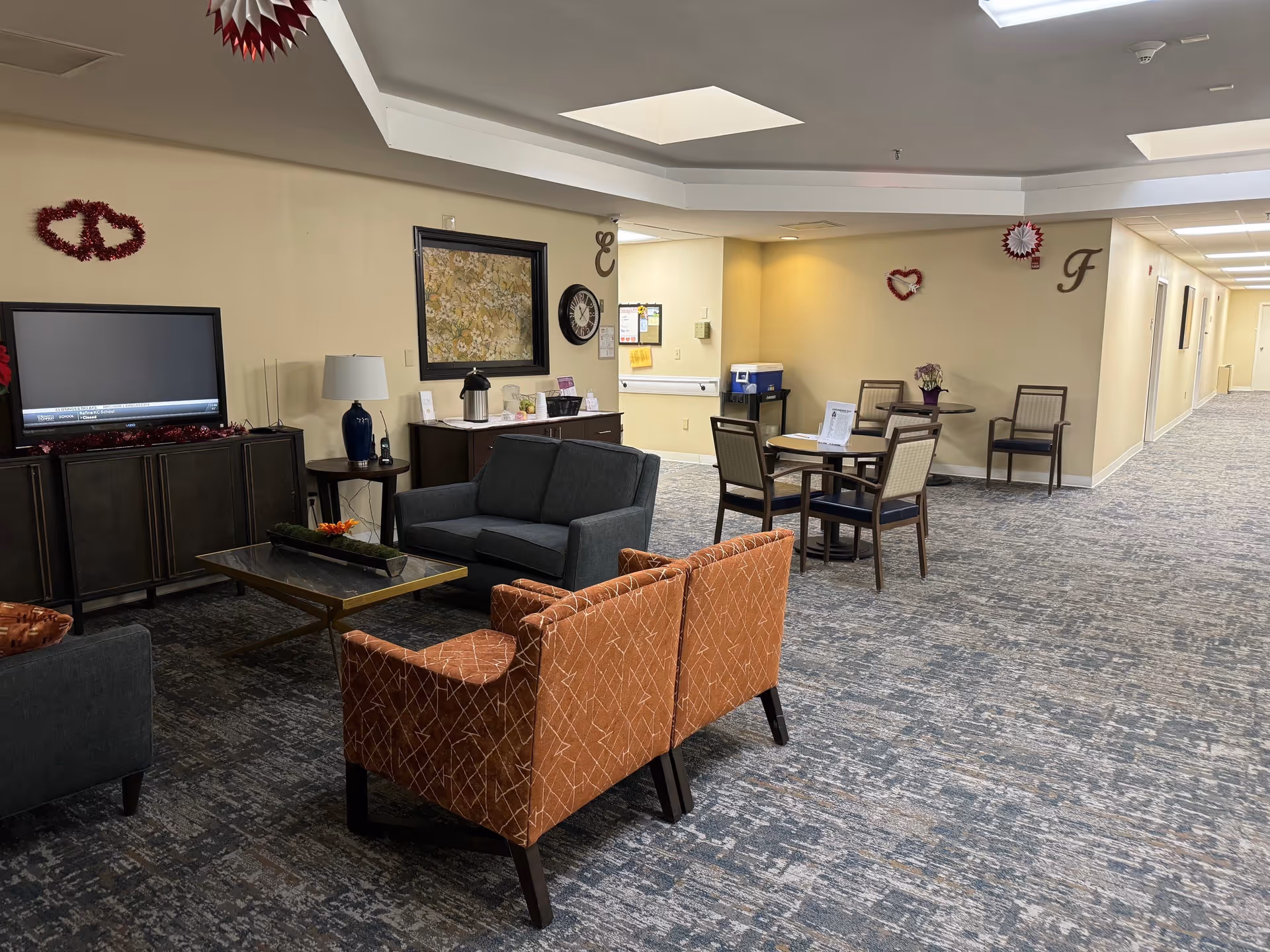 A senior living facility common area with a seating arrangement including a dark gray loveseat, two orange patterned armchairs, and a coffee table. There is a TV on a cabinet against the wall, a side table with a lamp, and a small dining table with four chairs. The walls are beige with some red heart-shaped decorations and letters 'E' and 'F'. The carpet is patterned in shades of gray and blue, and the ceiling has recessed lighting and skylights.