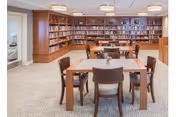 A well-lit library or reading room with wooden bookshelves filled with books along the back wall. There are several wooden tables and chairs arranged in the center of the room, with a small flower vase on one of the tables. The room has a carpeted floor and neutral-colored walls, creating a warm and inviting atmosphere.