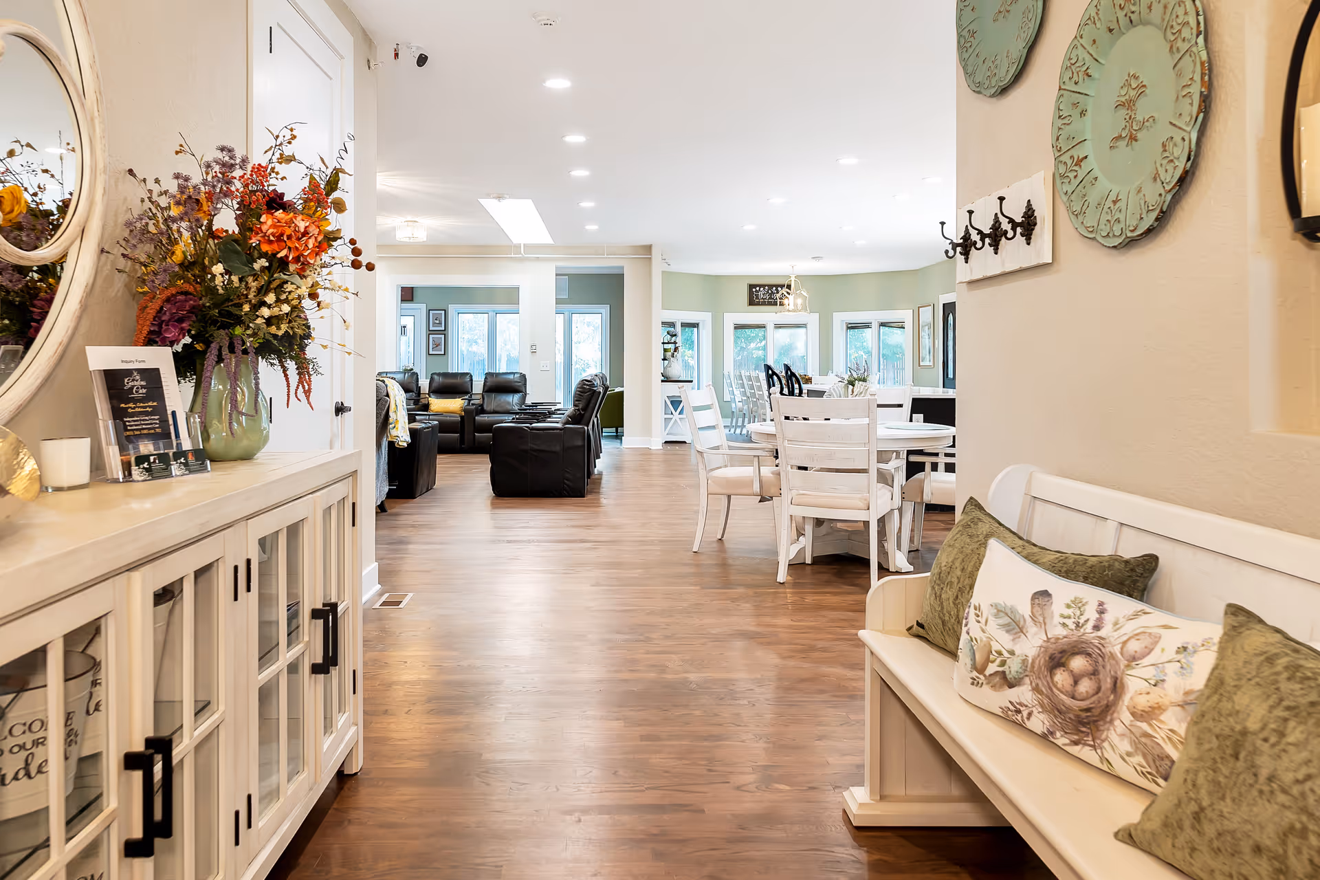 Interior view of a senior living facility showing a hallway with wooden flooring, a white bench with green and patterned pillows on the right, and a white cabinet with a vase of colorful flowers and a round mirror on the left. In the background, there is a dining area with white chairs and tables, and a living area with black leather recliners near large windows.