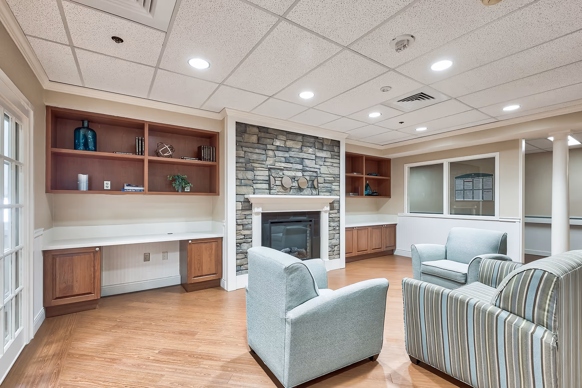 A cozy common area with a stone fireplace in the center, flanked by wooden shelves and cabinets. The room has light blue upholstered armchairs and a striped sofa arranged around the fireplace. The floor is wooden, and the ceiling has recessed lighting. There are windows and a glass door on the left side.