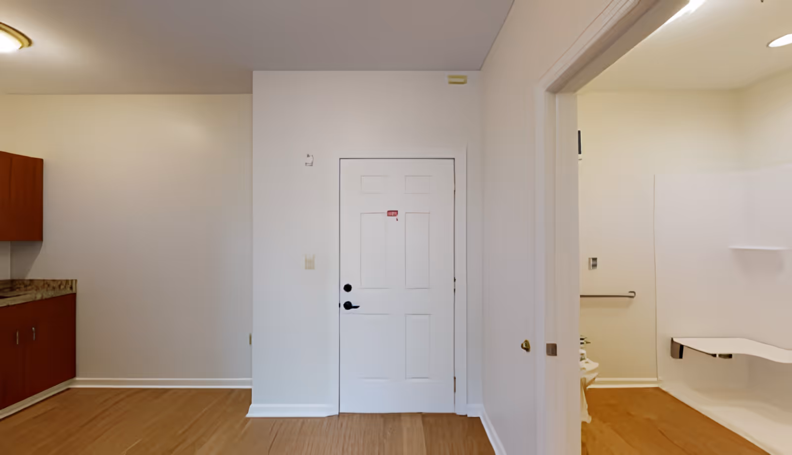 Interior view of a room with light-colored walls and wooden flooring. On the left side, there is a section of a kitchen counter with wooden cabinets and a granite countertop. In the center, there is a white door with a black handle. On the right side, an open doorway leads to a bathroom area with a white shower seat and grab bars.