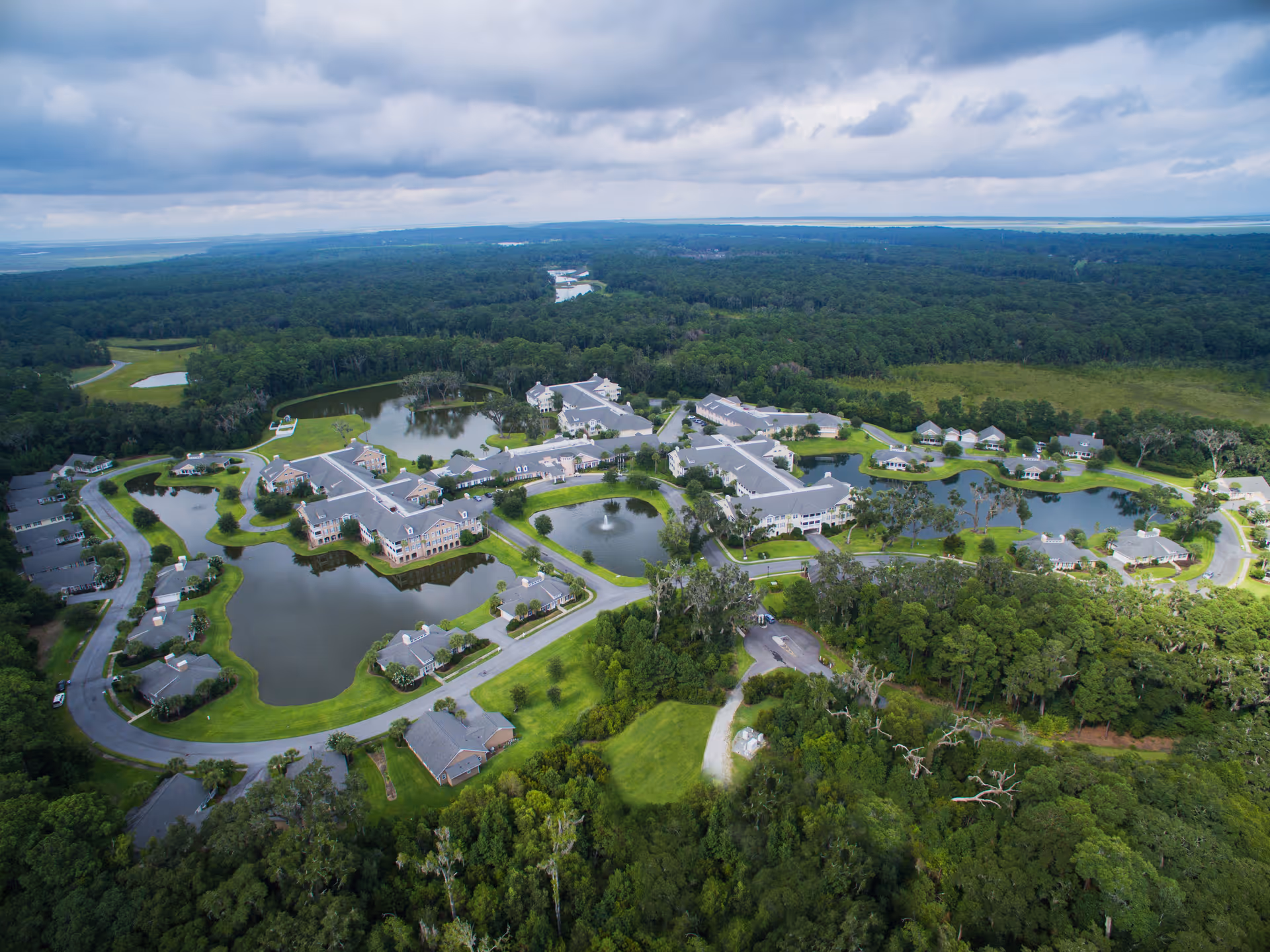 Aerial view of Marsh's Edge senior living facility surrounded by lush greenery and multiple ponds with fountains. The facility consists of several buildings connected by roads and pathways, set in a serene natural environment under a cloudy sky.