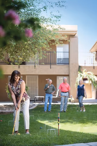 A group of four seniors playing croquet on a grassy lawn in front of a two-story building with balconies. One woman is preparing to hit a ball with a mallet while three others watch and smile in the background.