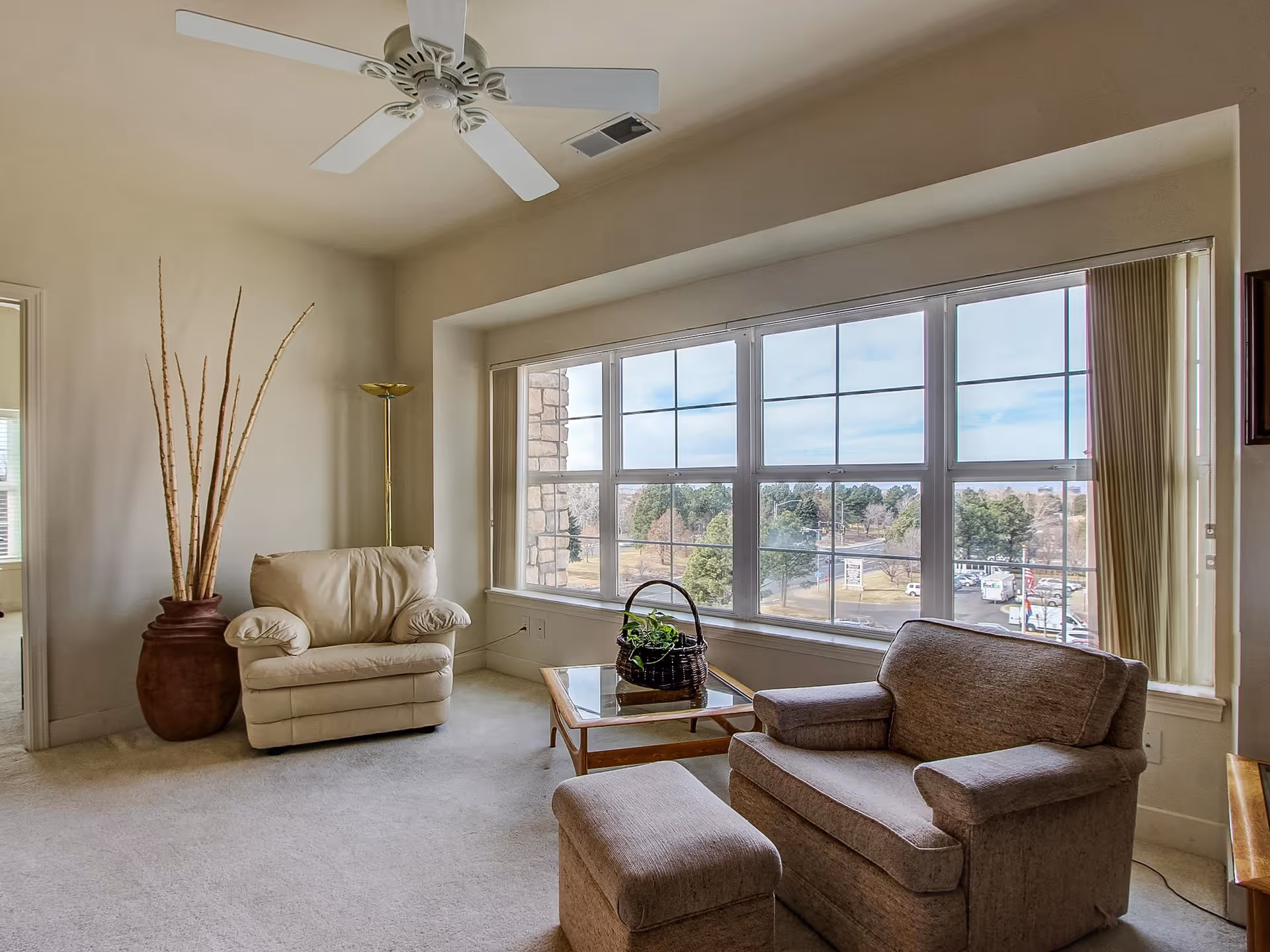 A cozy living room area with a large window showing an outdoor view. The room features a beige armchair, a brown upholstered armchair with matching ottoman, a glass-top coffee table with a basket plant, a tall floor lamp, and a large decorative vase with tall sticks. A ceiling fan is mounted above.