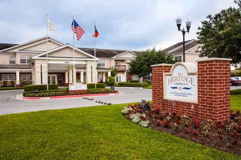 Front view of The Heritage Tomball Senior Living with entrance, flags, and landscaped sign.