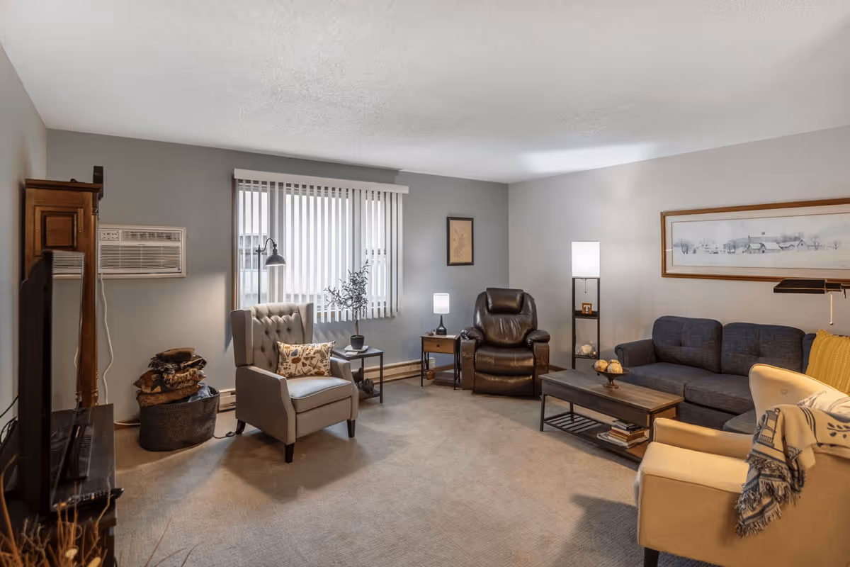 A cozy senior living room with a gray armchair, a brown leather recliner, a dark blue sofa, and a beige armchair. The room has a window with vertical blinds, a wooden coffee table with decorative items, a floor lamp, and a side table with a small lamp and a plant. The walls are painted light gray and decorated with framed artwork.